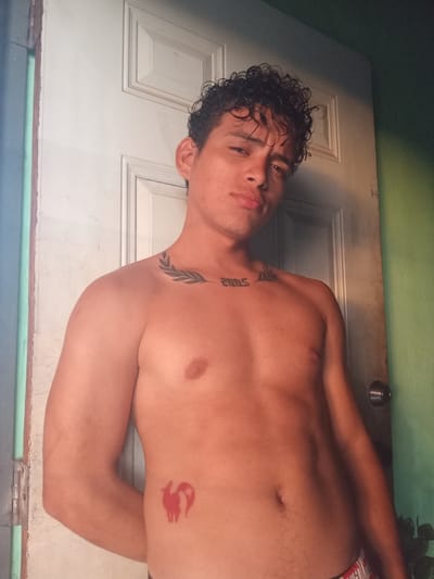 A young adult male, with curly dark hair, stands shirtless in front of a white door in a room with a green wall. He has tattoos on his chest and torso. One tattoo on his upper chest, near the collarbone, appears to be a laurel branch design with the number "2005" below it. Another tattoo on his right side, over the ribs, is a red silhouette of a fox-like creature. His arms are crossed behind his back. The environment suggests a casual, indoor setting, possibly a residence. The image does not provide any specific clues to its exact location within La Guardia, Venezuela, beyond it being an interior space.