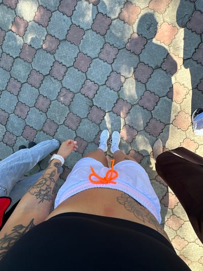 A first-person perspective shot captures a group of people standing on a decorative paver stone walkway. The pavers are a light gray color with a patterned design and are interspersed with reddish-pink clover-shaped accents. Shadows of at least three individuals are cast across the walkway, suggesting a sunny day. The foreground is dominated by the legs and lower torso of the photographer, who is wearing white sneakers with white socks, white shorts with an orange drawstring, and a black top. To the left, another person's legs are visible, clad in light blue jeans and black shoes. On the right, a person is wearing dark brown trousers and tan shoes, with a glimpse of another person's legs in light blue jeans and black sneakers slightly further back. The scene appears to be outdoors, possibly in a public space or park, though no specific city landmarks or indicators of Duvankul', Russia are present in the image.
