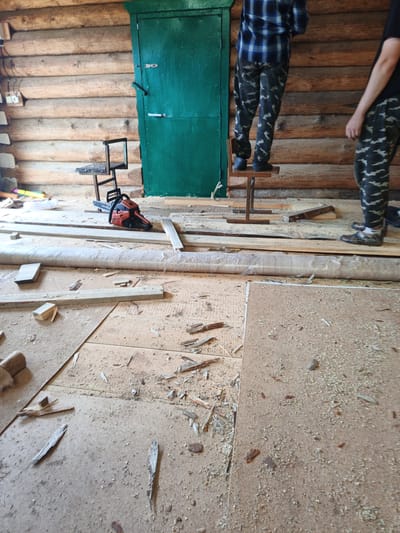 In an indoor, unfinished wooden structure, possibly a cabin or workshop, two individuals are engaged in some form of renovation or construction. The walls are constructed from horizontal logs. A green door is visible against the back wall. In the foreground, the floor is covered with debris, including wood shavings, broken pieces of wood, and what appears to be plywood or particleboard. Construction materials, such as planks of wood and a roll of material wrapped in plastic, are also scattered throughout the scene. A red and black chainsaw lies on the floor, suggesting it has been used or is about to be used. One person, wearing a blue plaid shirt and camouflage pants, stands on a small wooden stool, facing away from the camera. Another person, wearing a black t-shirt and camouflage pants, stands to the right, also facing away. The lighting suggests it is daytime, and the environment is dusty and appears to be in a state of disarray consistent with ongoing work. The location is identified as Islambaevo, Russia.