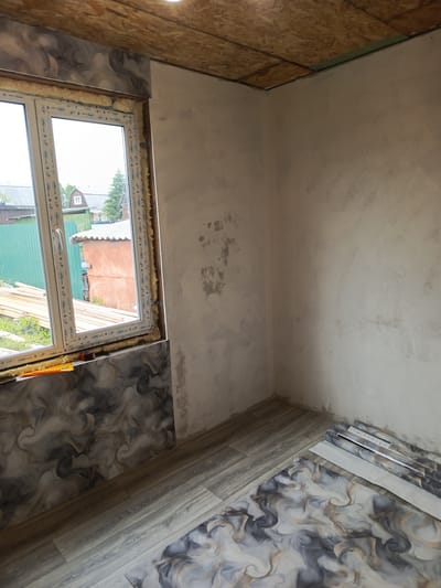 The image depicts an interior view of a room under renovation in Meget, Russia. The walls are plastered and appear unfinished, with some patchy areas. The ceiling is constructed from oriented strand board (OSB). A white uPVC window with double glazing is installed, framed by expanding foam insulation and decorative panels featuring a dark, swirling marble-like pattern. The floor is partially covered with laminate flooring and rolls of wallpaper, also with a similar marbled design. Outside the window, a residential backyard is visible, including a green fence, stacks of lumber, a small orange building, and houses. No people are present in the scene. The overall impression is of a room in the early stages of interior finishing.