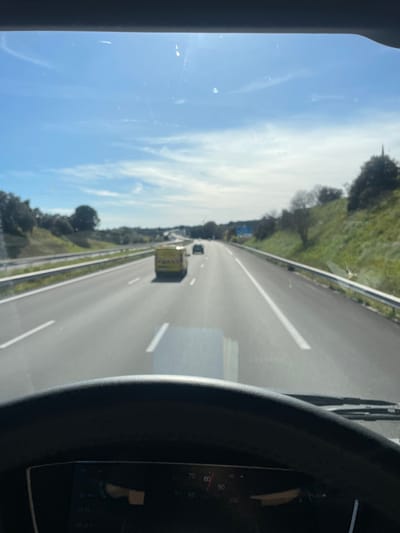 The image displays a daytime scene from inside a vehicle traveling on a multi-lane highway in Tavel, France. The top portion of a dark steering wheel and a dashboard speedometer, showing a speed of approximately 82 units (likely km/h), are present at the bottom of the frame. The asphalt road features white dashed lane markings and extends into the distance. A bright yellow utility van is positioned in the middle lane, moving away from the camera, with a dark passenger vehicle visible further ahead. Guardrails line the right side of the highway, beyond which are vegetated slopes covered with green grass and various trees and shrubs. The sky is clear blue with scattered thin white clouds. A blue road sign is partially visible on the distant right side of the highway.