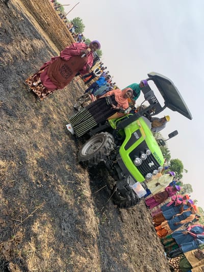 The scene is a gathering of people in Ringimawa, Nigeria, located at the coordinates 11.666 latitude, 8.414 longitude. There are several people present, both men and women. The people are standing in front of a lime green tractor, while others are seated on the tractor. The tractor is brand new. The people are dressed in various colorful clothing. The ground is covered in black burnt grass, possibly the aftermath of a recent fire. One person is holding a microphone and addressing the group. The overall atmosphere is one of celebration and excitement.