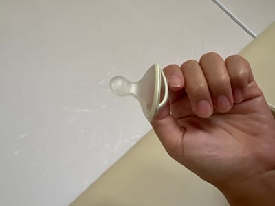 A close-up, overhead shot shows a hand holding a white baby pacifier against a light-colored, textured background. The hand, belonging to a person, is gently grasping the pacifier. The pacifier itself is made of clear silicone and has a white plastic shield. The background appears to be a wall or a surface with some subtle variations in lighting, suggesting an indoor setting. There are no prominent details about the time of day, weather, or specific location cues that would pinpoint a precise setting within Үшарал, Kazakhstan. The overall mood is neutral and focused on the object. There is no visible text in the image.