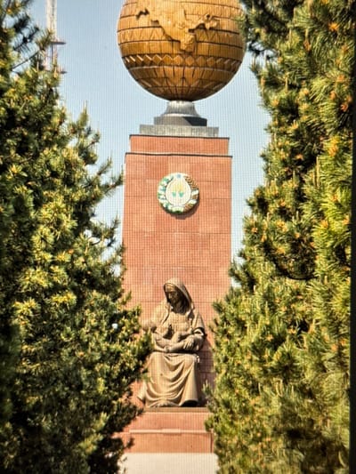 A tower appears surrounded by evergreen trees. The tower features a round, golden structure resembling a globe on one side, and a sculpture of a cloaked figure on the other. Centrally placed is a circular emblem with a green and white pattern. The tower itself is a light brown brick, creating a contrast with the natural greenery surrounding it. The image is taken from a low angle, emphasizing the height of the tower.
