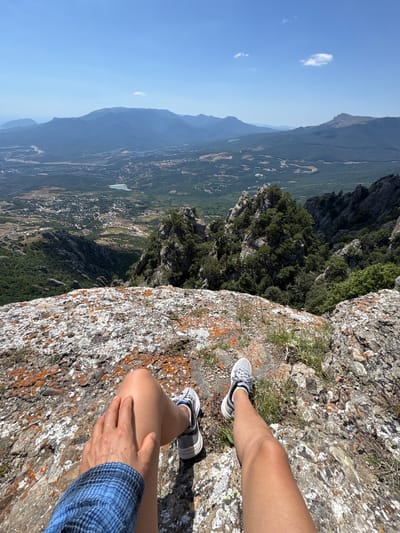 A first-person perspective shot shows two pairs of legs from the knees down, resting on a rocky cliff face. The legs are adorned with grey athletic shoes, one pair with white accents and the other with black. A hand with a blue plaid sleeve rests on the left leg. The background reveals a vast, verdant valley dotted with buildings and leading to a range of hazy blue mountains under a clear sky. The rocky surface in the foreground is textured with patches of orange and white lichen. The location is identified as the city of Luchistoe, Ukraine.