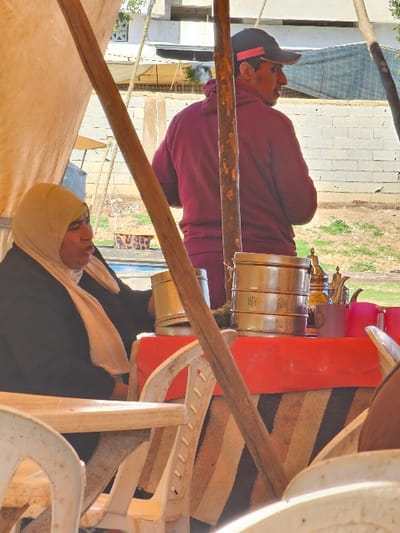 A man wearing a baseball cap and burgundy hoodie stands near a table of shiny metal objects. Another person, wearing a tan headscarf and black shirt, is seated. The scene is framed by wooden beams, suggesting a tent structure. Background elements include buildings, greenery, and stacked white plastic chairs, giving the impression of an outdoor market or event.