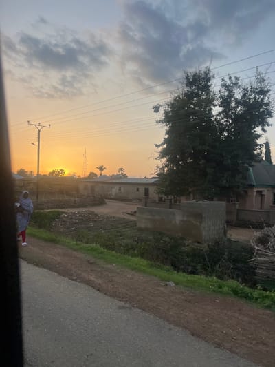 The image depicts a semi-rural or semi-urban scene in Ribina East, Nigeria, at either sunrise or sunset, characterized by a prominent orange-yellow sky on the left and center, transitioning to a lighter blue-grey with scattered clouds overhead.

In the immediate foreground, a dark vertical element partially obstructs the left side of the frame, suggesting the image was captured from within a vehicle. A grey, textured paved road or path occupies the lower left corner, diagonally extending upwards to the right. Adjacent to it, a reddish-brown dirt path runs parallel. To the right of the dirt path, a strip of green grass and vegetation is visible.

A person is observed walking on the dirt path in the left-midground, moving towards the right side of the frame. The individual wears a light-colored, full-length garment and head covering, bright red trousers, and white sandals. They appear to be carrying a light-colored bundle or possibly a small child close to their left side.

Beyond the grassy strip, the landscape consists of extensive unpaved brown earth, interspersed with areas of green vegetation, possibly small cultivated plots, and debris piles. Numerous single-story buildings, constructed from concrete blocks or similar materials, are scattered throughout the midground and background. Many appear weathered.

A utility pole with multiple overhead power lines and an attached street lamp stands prominently in the left-midground. To the right, a tall, slender metal tower, likely a telecommunications mast, is silhouetted against the bright sky. Various trees are present, including several distinctive palm trees in the mid-distance and denser, dark-leaved trees with bare upper branches on the right side of the frame. In the center-right midground, a partially constructed or dilapidated concrete block structure is visible, with a pile of dry sticks or wood to its immediate right. The setting or rising sun is a bright, intense orange orb in the left-center background, partially obscured by trees and distant structures.