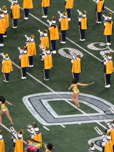 This image depicts a performance on an artificial turf football field within MetLife Stadium, East Rutherford, New Jersey, United States. The field features white yard lines and a prominent gray-and-white New York Jets "NY" logo, stylized as an oval. Approximately 20 individuals, identified as marching band members, are visible across the frame. They wear dark blue uniforms with yellow-gold capes, matching shakos adorned with plumes, and dark footwear, holding silver brass instruments in various performance stances. In the mid-right, a female performer in a metallic gold, long-sleeved leotard is captured mid-action, barefoot, holding a silver baton with both arms extended, one leg raised. Another performer in a similar gold costume is partially visible in the lower-left corner. A third performer, also in a gold costume, holds a multi-colored flag (red, purple, green, yellow) in the bottom-center. The performers are arranged in formations on the green turf, suggesting a coordinated group display.