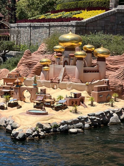 This image captures a meticulously crafted miniature scene resembling a desert oasis village, likely a themed attraction. The architectural style is reminiscent of Middle Eastern or Arabian designs, with sand-colored buildings, arched doorways, and numerous golden domes topping spires and structures. The foreground features a body of dark, rippling water, with rocky shorelines. Behind the village, sculpted red rock formations rise, creating a layered backdrop. Lush green vegetation and a vibrant, multi-colored floral display (red, yellow, and green) adorn the top of a stone wall in the upper portion of the image. The scene is bathed in bright, natural sunlight, indicating it's daytime. This setting is characteristic of Disneyland Park in Anaheim, California, specifically the "Aladdin's Oasis" or a similar Fantasyland attraction. There are no people or animals visibly depicted in the scene.