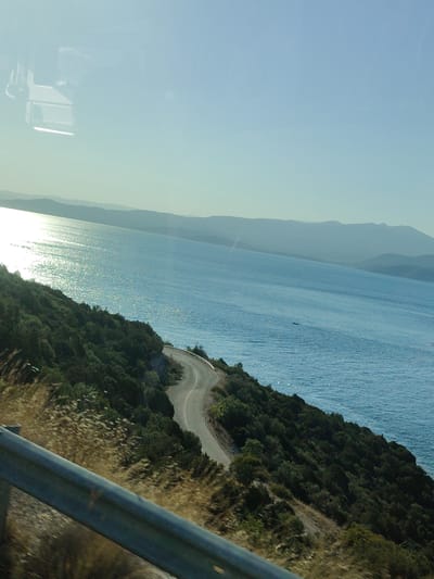 The image depicts a scenic coastal road winding through a green, hilly landscape overlooking a vast expanse of the Aegean Sea. The sun glints off the water, creating a shimmering effect. In the foreground, a metal guardrail runs along the edge of the road, suggesting a viewpoint from a moving vehicle. The road itself is asphalt, with a white line marking its edge. The vegetation is dense and appears to be a mix of trees and shrubs. In the distance, hazy mountains form a layered backdrop against the clear blue sky. The sky transitions from a pale blue near the horizon to a deeper blue overhead. The image is likely captured from a car driving along the coastal highway near the Municipal Unit of Pelasgia, Greece, known for its stunning sea views and winding roads. There are no people or animals visible in the frame.
