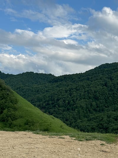 The image shows a vast landscape with a steep, tree-covered mountain dominating the right side of the frame. The mountain is covered in a dense, dark green forest. In front of the mountain, a lush green hillside slopes downwards, with patches of grass and scattered trees. To the far right of the image, there is a light-colored, uneven ground, possibly a dirt path or a field of dry vegetation.
The sky above is a mix of blue and white clouds, with the clouds appearing fluffy and scattered. The lighting suggests it is daytime, likely mid-morning or afternoon, with the sun illuminating the scene. The weather appears to be clear with some cloud cover.
There are no people or specific objects visible in the image, focusing solely on the natural landscape. The scene depicts a wild, natural environment, possibly in a mountainous region. The overall impression is one of natural beauty and expansiveness. The location is specified as Verkhnyaya Mara, Russia. There is no visible text in the image.