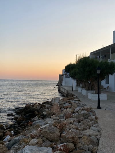 The image depicts a coastal scene in Apollonia, Greece, at dusk. The sky transitions from a light orange and pink near the horizon to a pale blue overhead. The sea, appearing calm and dark blue, occupies the left side of the frame, with a faint, distant dark shape, possibly a vessel, on the horizon. The immediate shoreline is rugged, composed of a dense accumulation of various-sized rocks in shades of grey, brown, and reddish-brown, extending from the water's edge to a concrete or paved walkway.
Along the right side of the walkway, a row of five neatly trimmed trees with dense green foliage are planted in white, square concrete planters. Four black, traditional-style streetlights with lantern tops are spaced between these trees. Behind the walkway, a series of multi-story, white-painted buildings with flat roofs are visible. One building features a covered overhang or balcony from which various articles of laundry are hanging to dry. To the far right, an outdoor seating area with two light-colored chairs and a small table is present on the paved surface adjacent to a building wall. Utility poles and power lines are visible above the buildings. No individuals are present.