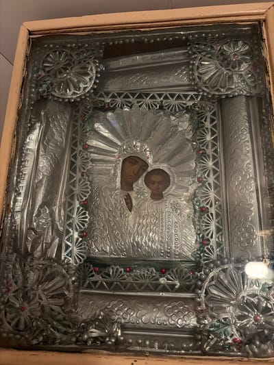 This is a close-up, eye-level shot of an ornate, silver-toned icon of the Virgin Mary and child Jesus, housed within a wooden frame and protected by glass. The icon features intricate embossed detailing, with the figures of Mary and Jesus depicted centrally, adorned with jeweled halos and ornate clothing. The surrounding metalwork is richly decorated with geometric patterns, floral motifs, and small red embellishments, resembling small beads or gems. The overall impression is one of religious significance and craftsmanship. There is no discernible indication of the city of Tambov, Russia, within the image itself.