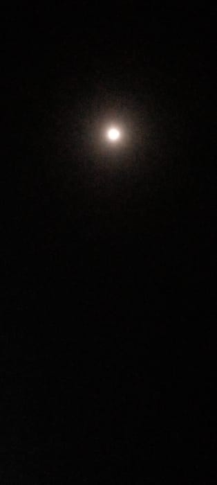 The image displays a bright, circular celestial body, identified as the moon, centered in the upper third of a dark, vertical frame, consistent with a night sky. The moon exhibits a soft, white luminescence with a diffused halo that gradually blends into the surrounding blackness. The background is uniformly dark, with faint, scattered, minute specks visible across the field, suggesting atmospheric particles or distant stars. No other identifiable celestial bodies, terrestrial features, man-made structures, people, objects, actions, or interactions are visible within the frame. The scene is located in Agunmo, Nigeria.