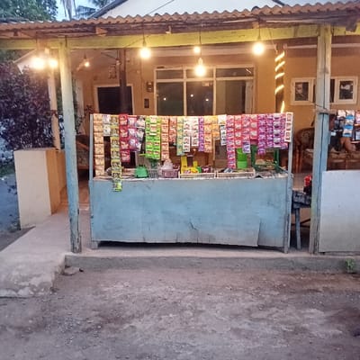 This image depicts a street vendor stall in Ketowan, Indonesia, illuminated during dusk or night. The foreground shows an uneven dirt and concrete ground leading to a raised concrete platform supporting the stall. The stall itself consists of a weathered, light-blue wooden or plywood counter. Numerous small, brightly colored packaged goods (sachets and snack bags) are suspended from strings stretched horizontally across the upper portion of the stall, forming a dense display. On the counter, a small green scale and other packaged items within plastic baskets are visible.

Above the stall, a corrugated metal roof, painted green and yellow, is supported by rough wooden posts. Several exposed incandescent light bulbs hang from this roof structure, providing the primary illumination for the scene. Behind the stall, a light-colored (tan or beige) building with multiple windows and a door is visible; interior lighting is active. To the far right within the building, an ornate chair is discernible, and a person's lower leg and arm are faintly visible, suggesting an individual is seated. Decorative string lights ascend a vertical element on the building's facade to the right. To the left of the stall, a dark-leaved bush is situated behind a low, light-colored concrete planter.