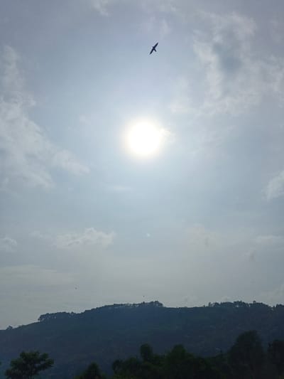 A bright sun dominates the sky, filtering through a thin layer of clouds. Above the sun, a bird soars. Below, a distant, silhouetted mountain range fades into the horizon. The landscape features a dark, forested ridge in the foreground, adding depth to the scene. The hazy atmosphere softens the colors and contrasts, creating a serene, nature-filled image.