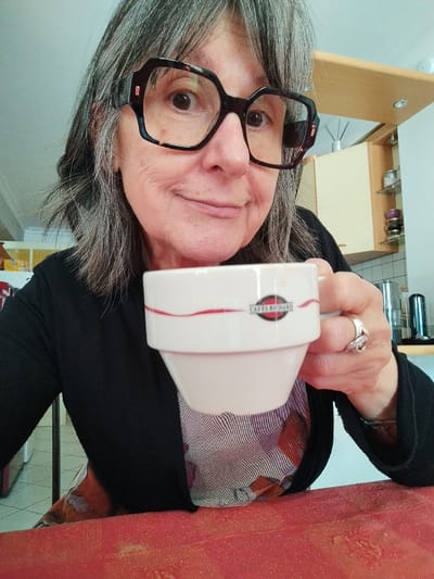 The image depicts an older woman in a private residence located in Montélimar, France. She is positioned centrally, looking directly at the camera with a subtle smile. Her gray hair is shoulder-length, and she wears large, dark-framed eyeglasses. She is dressed in a black long-sleeved cardigan over a multi-colored patterned garment. Her left hand holds a white ceramic cup with a red curved line design and a "CAFÉS RICHARD" logo in a black and red oval. A small brown stain is visible near the rim of the cup. The immediate foreground features a reddish-orange, textured surface, possibly a tabletop. The background reveals an indoor domestic setting, specifically a kitchen area, with light wooden upper cabinets, a white tiled backsplash, and various kitchen items including glass jars on shelves, a reed diffuser, and a silver cylindrical container.