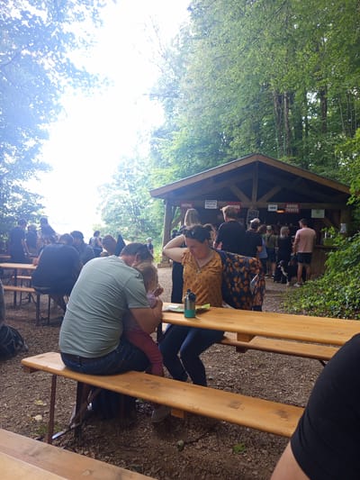 In the city of Azannes-et-Soumazannes, France, amidst a wooded area, a gathering of people are seen at an outdoor wooden kiosk. The kiosk, constructed from wood, displays signage for "CRÊPES," "Glace de Meuse!," and "CAISSE TICKETS." Several individuals are lined up at the kiosk, interacting with staff. In the foreground, a man and a woman are seated at a wooden picnic table. The man is holding a young child on his lap. Another person, partially visible, is also seated at the table. The ground is covered with gravel and fallen leaves, and the surrounding environment is lush with trees and foliage. The overall scene depicts a casual outdoor gathering at a food and ticket vendor.