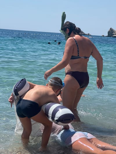 The image shows a group of people on a beach, with the water lapping at their feet. A woman in a bikini is bent over, wearing a snorkel and mask. Next to her, a young boy in swim trunks is sitting in the water, also wearing goggles and a snorkel. He has inflatable armbands with blue and white stripes. Another person's legs are visible on the right side of the image, wearing a light blue and white striped shirt. The water is clear and shallow, with sunlight glinting off its surface. In the background, the calm, blue sea stretches out to the horizon, with a rocky outcrop visible on the left. The sky is clear and bright, suggesting it is a sunny day, likely in the morning or afternoon. The scene appears relaxed and joyful, indicative of a holiday or recreational outing at the beach. The overall atmosphere is summery and peaceful. There is no visible text in the image.