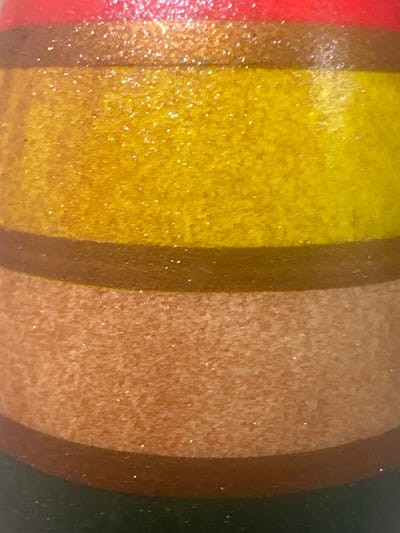 This is a close-up, detailed, abstract shot of a surface with horizontal stripes of color. The top stripe is red, followed by a thin brown stripe, then a wide yellow stripe with glitter, another thin brown stripe, and a wide tan stripe with glitter. The bottom of the image shows a dark green stripe. The image does not depict any elements that would allow for the identification of a specific location in Letterkenny, Ireland.