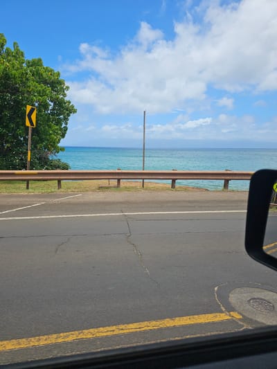 The image shows a scenic view of the ocean from a roadside in Napili, Maui, Hawaii, USA. In the foreground, a portion of a car's side mirror and the asphalt road with yellow lane markings are visible. A guardrail made of rusty metal runs along the edge of the road, separating it from a grassy area and then the turquoise ocean. A large green tree with lush foliage is on the left, with a yellow directional sign indicating a curve. The sky is a vibrant blue with scattered white clouds. In the distance, the ocean stretches out to meet the horizon, with a faint outline of another island visible. A tall, thin pole stands near the center of the frame. The scene appears to be captured on a clear, sunny day.