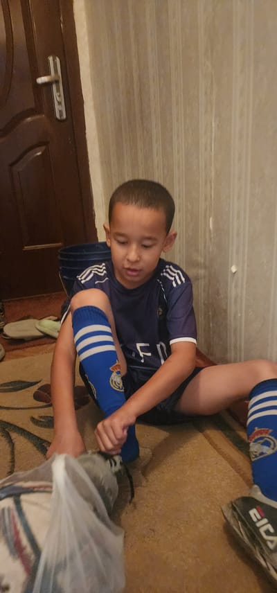 A young boy wearing a blue and white Real Madrid football jersey and blue socks with white stripes is sitting on a patterned rug indoors, attempting to put on a soccer cleat. He is looking down at his feet, and his hands are occupied with the shoe. To his left, a blue bucket is visible, and behind him, a brown door with a silver handle and a textured, light-colored wall are present. The overall environment suggests a residential setting within the city of Türkmenbaşy, Turkmenistan.