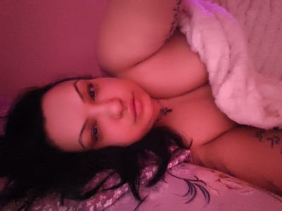 The media file shows a close-up, low-angle shot of a person lying down. The subject has dark hair and is partially covered by a white fluffy blanket. The lighting is dim and casts a pinkish hue over the scene. The person's face is visible, with one eye looking towards the camera. The background appears to be a bed with a patterned sheet. There is a tattoo visible on the person's arm. The overall impression is one of intimacy and relaxation.