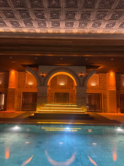The image shows a building exterior in Ürgüp, Turkey. The building features a decorative facade with carved stone panels on the left and a stone structure with arches in the center. The arches are illuminated with warm light. To the right is a swimming pool reflecting the building's lights. The scene is captured at night, and the lighting creates a warm, inviting atmosphere.