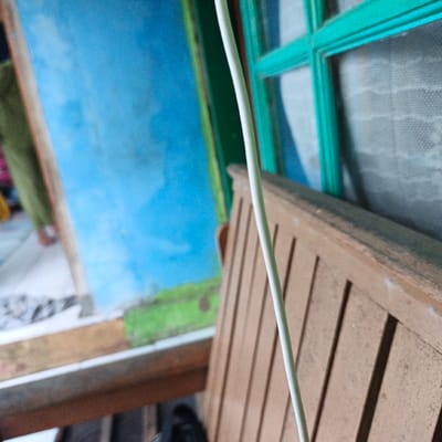 The image shows a wooden bench positioned in front of a window with a green frame and a white wire running vertically in front of the bench. The window is partially obscured by a white mesh screen. The bench is made of slats and has a light brown color. The wall behind the bench and window is painted a light blue color with green paint near the bottom. The bench appears to be outside, as there is a blur of green vegetation visible near the bottom. The image was taken at a location in Pameungpeuk, Indonesia, with coordinates -7.641 latitude and 107.728 longitude.