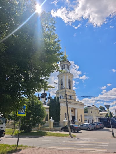 In Chekhov, Russia, under a bright blue sky dotted with fluffy white clouds, a yellow church with a tall bell tower stands prominently. The sun's rays streak through the leaves of a large, dark green tree in the foreground, partially obscuring the view of the church. A pedestrian crossing sign, a blue and yellow diamond shape with a depiction of people crossing, is visible on the left side of the image. Several cars are parked in front of the church, and a white truck is partially visible on the far left. The street in front of the church has a marked pedestrian crossing. The church has multiple dark domes and a golden cross atop the bell tower. Electrical wires and a lamppost are also visible near the church.