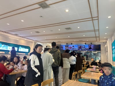 A bustling fast-food restaurant setting with a long line of customers awaiting service. A digital menu board displays various meal options above the counter. Patrons are seated at tables, enjoying their meals. The restaurant features a bright interior with wooden accents and modern lighting, conveying a casual dining atmosphere. A young boy in a patterned jacket sits at a table in the foreground.