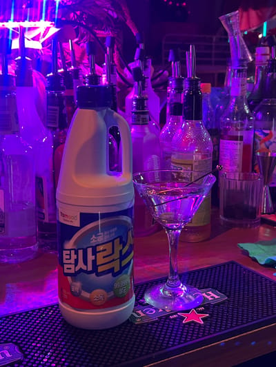 The scene depicts a bar counter under purple and blue neon lighting in 마포구, South Korea. A white plastic bottle of TamSok brand bleach, labeled in Korean as "탐락스" and indicating 99.999% sterilization, occupies the left foreground, standing on a black rubber bar mat. To its right, a clear martini glass containing a transparent liquid and a black cocktail pick rests on the same bar mat, positioned over a green Heineken coaster featuring a red star. Behind these items, numerous clear and colored liquor and syrup bottles, many equipped with metal pour spouts, are arranged on the wooden counter. A "MONIN" brand syrup bottle is visible among the collection. A silver bar jigger is partially visible on the far right. No individuals are present in the image.