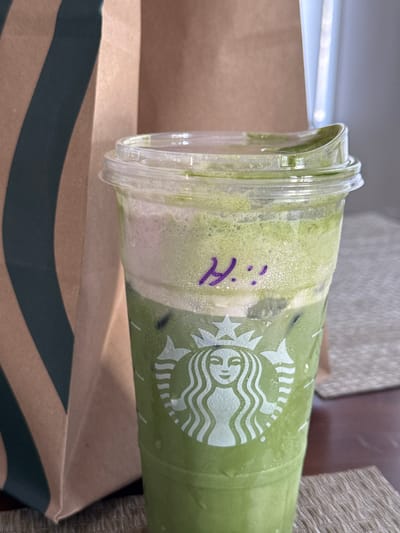 A slightly tilted view captures a Starbucks cup on top of a Starbucks bag in what appears to be an indoor setting. The cup is filled with a green drink that appears frothy with condensation on the cup. The Starbucks logo is visible. "Hi" is written on the cup in purple marker. The bag is brown with a dark green stripe. The setting is likely somewhere in Chula Vista, United States of America.