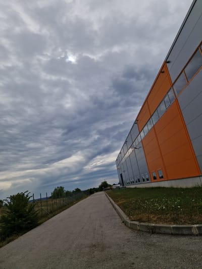 The image shows a modern industrial building with an orange and gray facade, situated in the city of Bahovitsa, Bulgaria. The building appears to be a warehouse or factory, with multiple windows and a large, flat roof. A paved road runs alongside the building, leading away from the viewer. To the left of the road, there is a chain-link fence and some green bushes. The sky is overcast with dramatic, textured clouds, and the sun is partially visible through a break in the clouds. The overall scene suggests an industrial area on the outskirts of the city. There are no people or vehicles visible in the image.