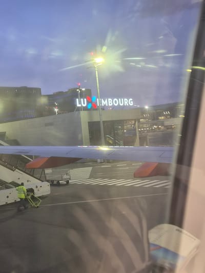 The image shows a view from inside an airplane at Luxembourg Airport (LUX) in Niederanven, Luxembourg. The wing of the airplane is visible in the foreground, and ground crew is working around the airplane's loading apparatus. The airport building is visible with "LUXEMBOURG" in large vertical block letters on its facade. Further in the background are buildings in the city. The view is likely through an airplane window with reflections.