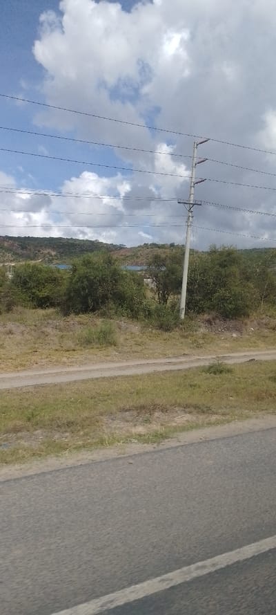 The image depicts a semi-arid, open landscape in Kinanie ward, Kenya, viewed from a low angle, likely from a moving vehicle. In the immediate foreground, the upper portion of an asphalt road is visible, featuring a faded white solid line on its right edge. Directly beyond the road, a dirt track runs parallel to it, bordered by dry grass and scattered low-lying scrub bushes. To the right mid-ground, a tall, slender utility pole, likely concrete, stands upright, supporting multiple horizontal power lines connected via reddish-brown ceramic insulators. These power lines extend horizontally across the upper middle section of the frame. The background reveals a series of undulating hills covered in sparse, dry vegetation and exposed reddish-brown earth. Distant blueish structures or objects are faintly discernible on the far hills. The sky above is partially cloudy, with prominent white and grey cumulus clouds interspersed with patches of bright blue sky. No individuals, animals, or active interactions are present within the frame.