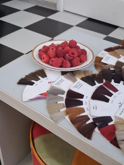In the city of Tuzdybastau, Kazakhstan, a close-up, top-down shot reveals a white table surface adjacent to a black and white checkered tiled floor. On the table sits a white porcelain bowl filled with fresh raspberries. To the right of the bowl are several fan-shaped color swatches, likely for hair dye, arranged in a semi-circular pattern. Some swatches display hair strands in various shades, while others have printed labels indicating colors and numbers such as "Intense Fashion," "Gold Beige Gold Copper," "Natural," "Copper Cold Mahogany Chocolate," "Red Violet," and numerical codes like "5.00," "6.00," "7.00," "3.8," "4.89," "5.56," "8.43," "4.55," and "8.77x." Underneath the table, a glimpse of a red and green object, possibly a bag or cushion, is visible within a shelf or recess.
