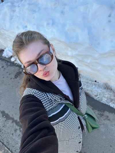 A forensic description of the scene in Новое Шигалеево, Russia:
An outdoor, high-angle selfie captures a female individual from the chest up, posing with pursed lips. The subject wears oversized brown-framed sunglasses with reflective lenses, partially obscuring her eyes. Her light brown hair is pulled back. She is dressed in a dark brown jacket featuring a prominent black and white houndstooth pattern across the torso, with solid dark brown sleeves and collar, over a light pink t-shirt. A green shoulder bag is worn across her body, visible on her right side. The background consists of a large accumulation of snow, exhibiting patches of melting and dirt, indicating cold weather conditions. A strip of dark grey pavement or asphalt is visible along the lower left edge of the frame. The scene is brightly lit by natural sunlight.
