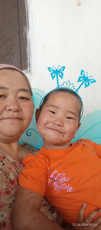 This image depicts a close-up, front-facing selfie of an adult and a young child, taken outdoors against a white wall with a dark-colored wooden element on the left. The adult, on the left, wears a pink headscarf and a floral-patterned top. The child, on the right, is adorned with blue butterfly wings and a headband featuring two blue butterflies and a small tiara. The child's face has red markings, resembling paint or stains, scattered across the cheeks and nose. The child wears an orange t-shirt with a pink floral design. The background consists of a textured white wall, with a darker, possibly wooden, vertical surface on the far left. The timestamp "25.06.2026 07:55" is visible in the bottom right corner. The scene appears to be in Chon-Kara, Kyrgyzstan.