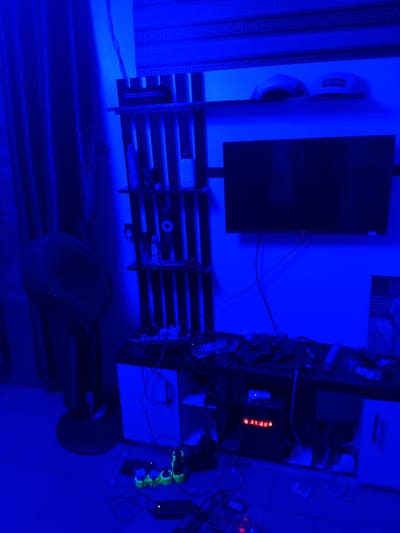 The image shows a room lit with blue ambient lighting. In the foreground, on the left, is a black oscillating fan with its base on the floor. To the right of the fan is a tall, dark shelving unit with multiple shelves. Various items are placed on these shelves, including what appear to be bottles or containers and possibly electronic devices. Above the shelves, a horizontal surface displays two baseball caps, one with a "Supreme" logo. A flat-screen television is mounted on the wall above this surface. Below the television, a black media console unit is visible, with several objects and electronic accessories scattered on top and within its compartments. A tangle of wires and cables is present around the media console and on the floor. On the floor in the lower right portion of the image, several power outlets with green glowing accents are visible, along with additional electronic devices and cables. The overall impression is of a personal entertainment or gaming space, likely a bedroom or living area. The specific location in Ogbogbo, Nigeria is not discernible from the visual information provided in the image.