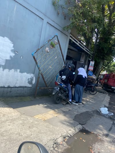 The scene is a sidewalk in the city of Kecamatan Dayeuhkolot, Indonesia. It is located at coordinates -6.963, 107.594. Two people are standing on the sidewalk. One is a man wearing a black jacket with the word "NIKE" on the back, a black helmet, and blue jeans. He is standing next to a black and blue motorcycle with a license plate that reads "4547 P 0635536". The other is a woman wearing a white hijab and a blue dress. They are both looking at each other. There is a large, metal trellis with plants growing in it, leaning against a grey building. The sidewalk is cracked and uneven, with a puddle of water in the middle. There is a green tree growing in the background, and a red truck parked on the side of the road. The truck is partially obscured by the tree and the man. There is also a black motorcycle parked on the sidewalk, behind the man. There is a white plastic bag on the ground next to the puddle.