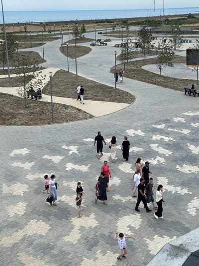 The image shows a paved walkway with a patterned surface, likely in a park or recreational area, near the coast. The sea is visible in the background under a cloudy sky. Several people are walking on the paved area. In the foreground, a young boy in a patterned t-shirt and shorts is in motion, appearing to throw something. A woman with sunglasses and a light blue skirt is walking with two children, one in a pink shirt and the other in a dark dress. Further back, a group of people are walking together, including a man in a black t-shirt and shorts, a woman in a long floral dress, a woman in a black dress, and other individuals. There are also benches and lampposts visible along the pathway. The area seems to be recently developed, with some areas of dry grass and young trees.