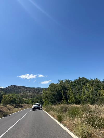 A bright, sunny day with a clear blue sky over a paved road that curves through a rural landscape. Two white cars are driving on the road, one directly behind the other, heading away from the viewer. The road is bordered by dry, tall grass and brush on the right side, and a mix of green trees and scrubby vegetation on the left. In the background, rolling hills covered in trees and rocks rise towards the horizon. A few scattered white clouds are visible in the sky. The image captures a scenic drive through a natural environment. Based on the context provided, this scene is located in the vicinity of Slivnitsa, Bulgaria.