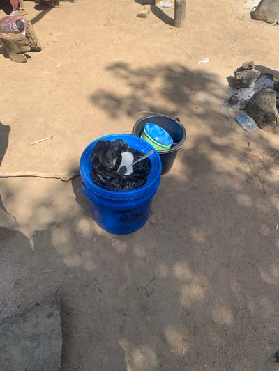 The image depicts an outdoor scene on a dirt ground, possibly in Project, Nigeria. In the foreground, a bright blue bucket is filled with black plastic bags and some white material. To the right of the blue bucket, a black bucket contains two colorful plastic bowls, one blue and one green. Scattered around the ground are rocks, a piece of wood, and some debris including plastic. Shadows from trees or structures are cast across the ground. The overall impression is one of a functional, outdoor workspace or living area, with evidence of daily activities such as the use of buckets for storage or carrying.