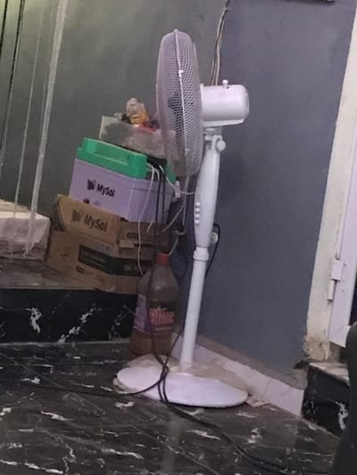 A white standing fan with a cage leans towards the left, partially obscuring a messy collection of items. These items include a green-lidded MySol battery box, a cardboard box with "MySol" printed on it, a plastic bottle with a red cap, and miscellaneous items wrapped in plastic. The background features a gray wall, a white window frame, and a marble-patterned floor. Electrical wires snake behind the fan.