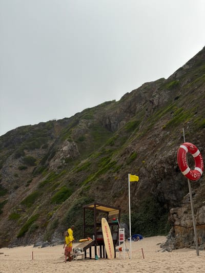 A wide-angle, eye-level shot captures a coastal scene in Colares, Portugal, under a hazy sky. A rugged, green-and-brown hillside dominates the background, its steep incline covered in sparse vegetation and rocky outcrops. In the foreground, a sandy beach stretches out, leading to the base of the hill.

Several elements of a beach rescue station are present. A wooden lifeguard tower with a yellow awning stands near the center, equipped with a surfboard leaning against its side. Next to the tower, a yellow flag waves from a pole. A red and white life preserver ring hangs from a metal pole on the right side of the frame, displaying Portuguese text and a phone number.

Two lifeguards, dressed in yellow shirts and shorts, are visible on the sand. One stands near the lifeguard tower, while the other walks away from the camera. A small blue tent is set up further back on the beach, where two individuals are seated. In the lower part of the image, a red rope barrier is partially visible. The overall scene conveys a sense of preparedness for beach safety in a natural, coastal environment.