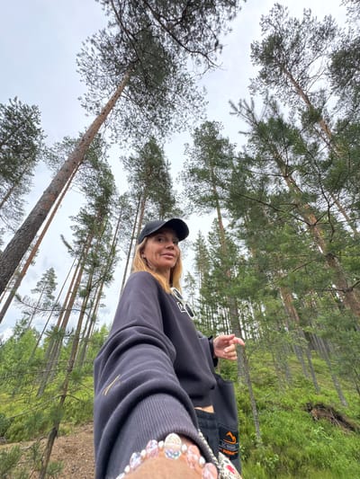 A low-angle, wide shot captures a woman with blonde hair and fair skin standing in a pine forest. She is wearing a black baseball cap, a dark grey sweatshirt, and dark pants with an orange logo on the right leg. Her left arm is extended towards the camera, and her left hand is visible, adorned with a bracelet of colorful beads. She is looking slightly to her right, with her mouth open. The background is dominated by tall, slender pine trees with rough bark, reaching towards a cloudy sky. The forest floor is covered in green undergrowth and pine needles. The scene appears to be in the city of Peski, Russia.
