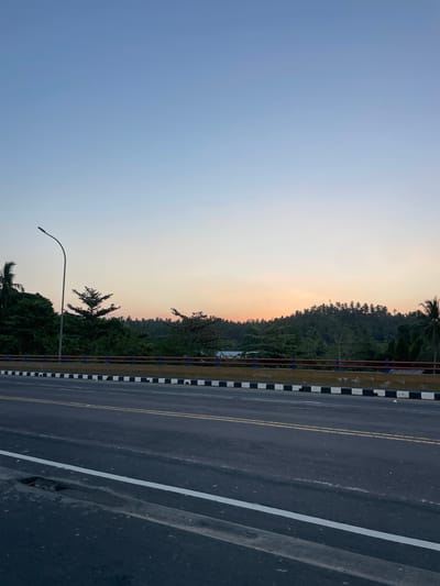 The image captures a wide shot of a road and a tree-lined hillside at sunset. The foreground is dominated by a dark asphalt road with double yellow lines indicating traffic lanes and a white line marking the edge of the road. A drain cover is visible on the left side of the road. A concrete barrier with black and white checkered patterns and a red and blue railing separates the road from the embankment.

Beyond the barrier, a dense canopy of various trees, including palm trees, covers a gently sloping hillside. The sky above is a gradient of pale blue at the top, transitioning to a soft orange and pink hue near the horizon, indicating the setting sun. A single lamppost stands on the left side of the road, its curved arm extending over the scene. The overall impression is of a tranquil evening scene on a bridge or elevated roadway in Maumbi, Indonesia.
