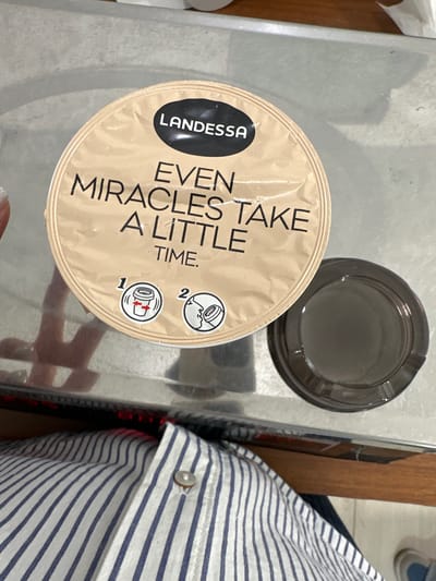 The scene shows a Landessa coffee capsule and a grey lid on a metallic surface. Part of a person wearing a blue and white striped shirt is visible to the right. The setting is not explicitly identified as Agarak, Armenia, nor is any location information provided. There is no evidence of actions or interactions in the image.