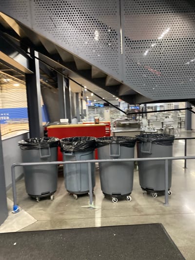 Four large, gray, wheeled trash cans with black liners are lined up next to a metal railing. The trash cans are positioned on a concrete floor. Behind the trash cans, a red counter is visible, and further in the background, tables and chairs are arranged in what appears to be a dining or seating area. Above, a metal staircase with a perforated panel runs diagonally across the frame. The environment suggests an indoor public space, possibly a recreation center or a venue in Virginia Beach, Virginia.